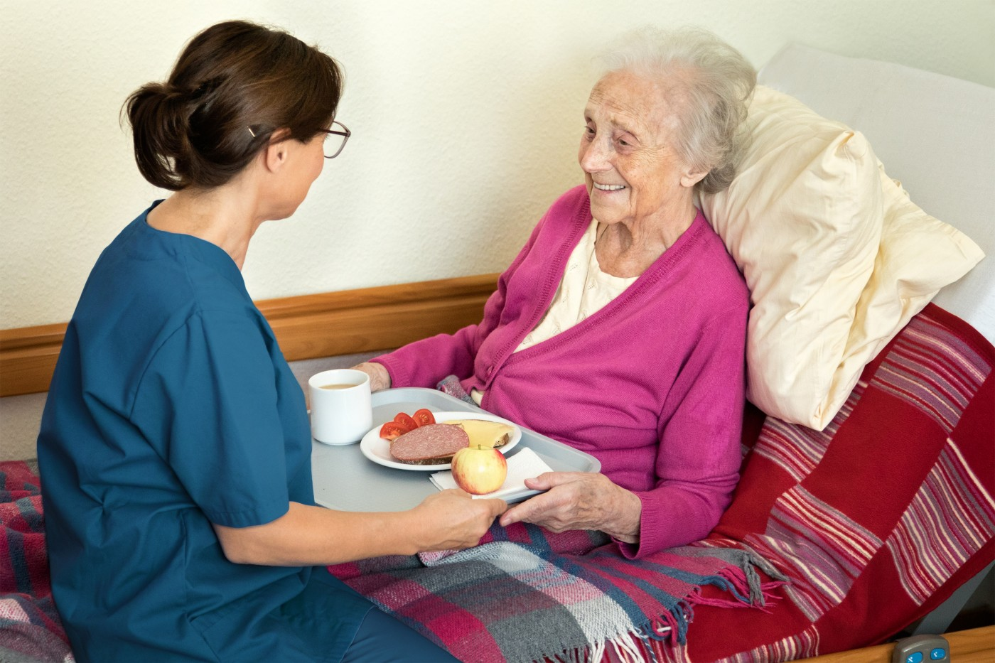 Caregiver in blue scrubs serving a meal to a smiling elderly woman in a pink cardigan who is comfortably seated with pillows and a colorful blanket